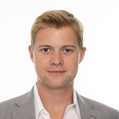A young man with short blonde hair wearing a light gray suit jacket and a white shirt, facing forward and smiling slightly, on a plain white background.