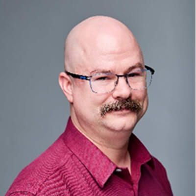 A middle-aged man with a bald head, glasses, and a mustache is wearing a maroon collared shirt and smiling slightly against a plain gray background.