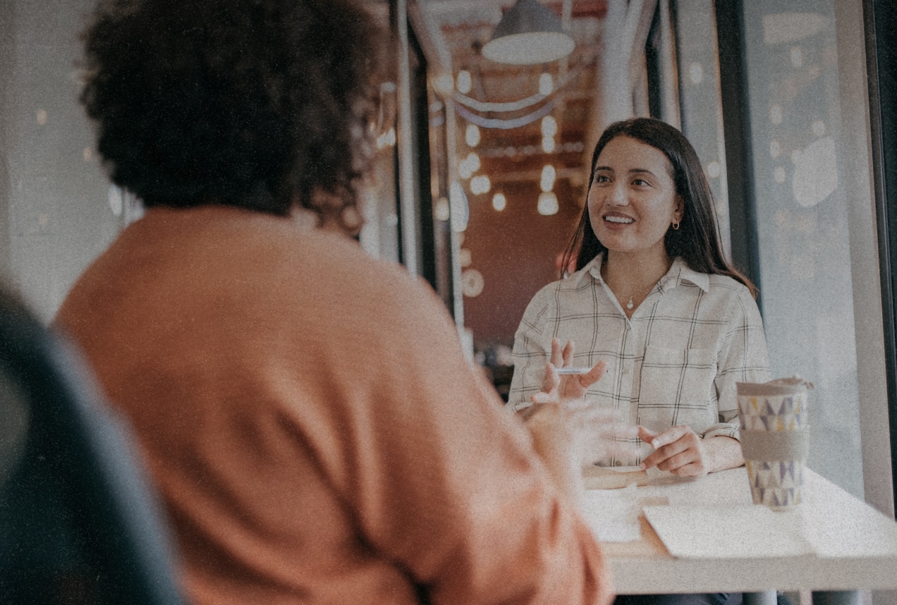 Two women sit across from each other at a table in a cozy café, engaged in conversation. One woman smiles while listening, and a coffee cup sits on the table between them. Warm lights hang in the background.