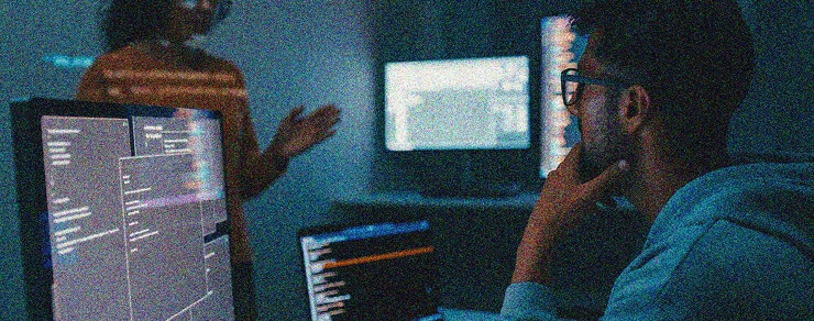 A person wearing glasses sits at a computer with code on the screen, focusing intently on Azure Sentinel Implementation. Another person stands in the background, gesturing toward a monitor in a dimly lit room filled with technology.