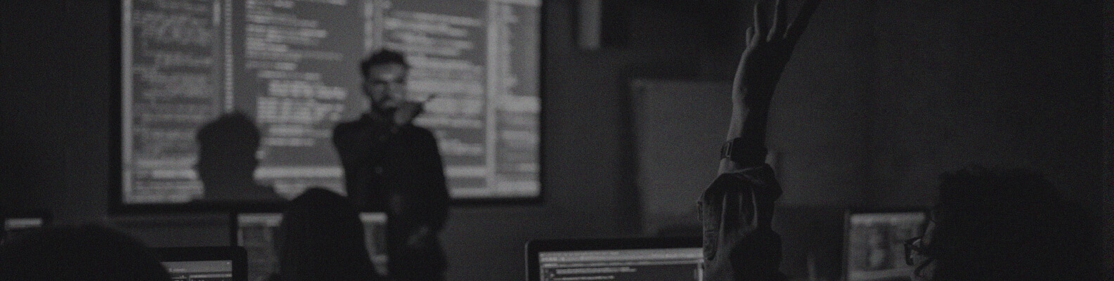 A person stands in front of a classroom with computer screens and a large projection of Azure Sentinel Implementation code, while a student in the foreground raises their hand to ask a question.