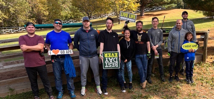 A group of ten people, including a child, stand outdoors in front of a wooden fence holding fun signs with phrases like “I ♥ Go Ape,” “Best Day Ever,” and “I Went Ape.” Trees and grass are visible in the background.