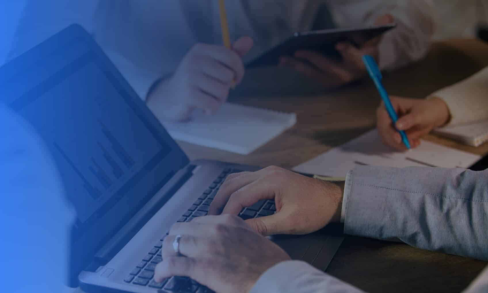 Several people sit around a table in a bright office, working on laptops and using tablets and smartphones. Graphs on a laptop screen suggest an Azure Sentinel Implementation is being discussed in this collaborative business meeting.