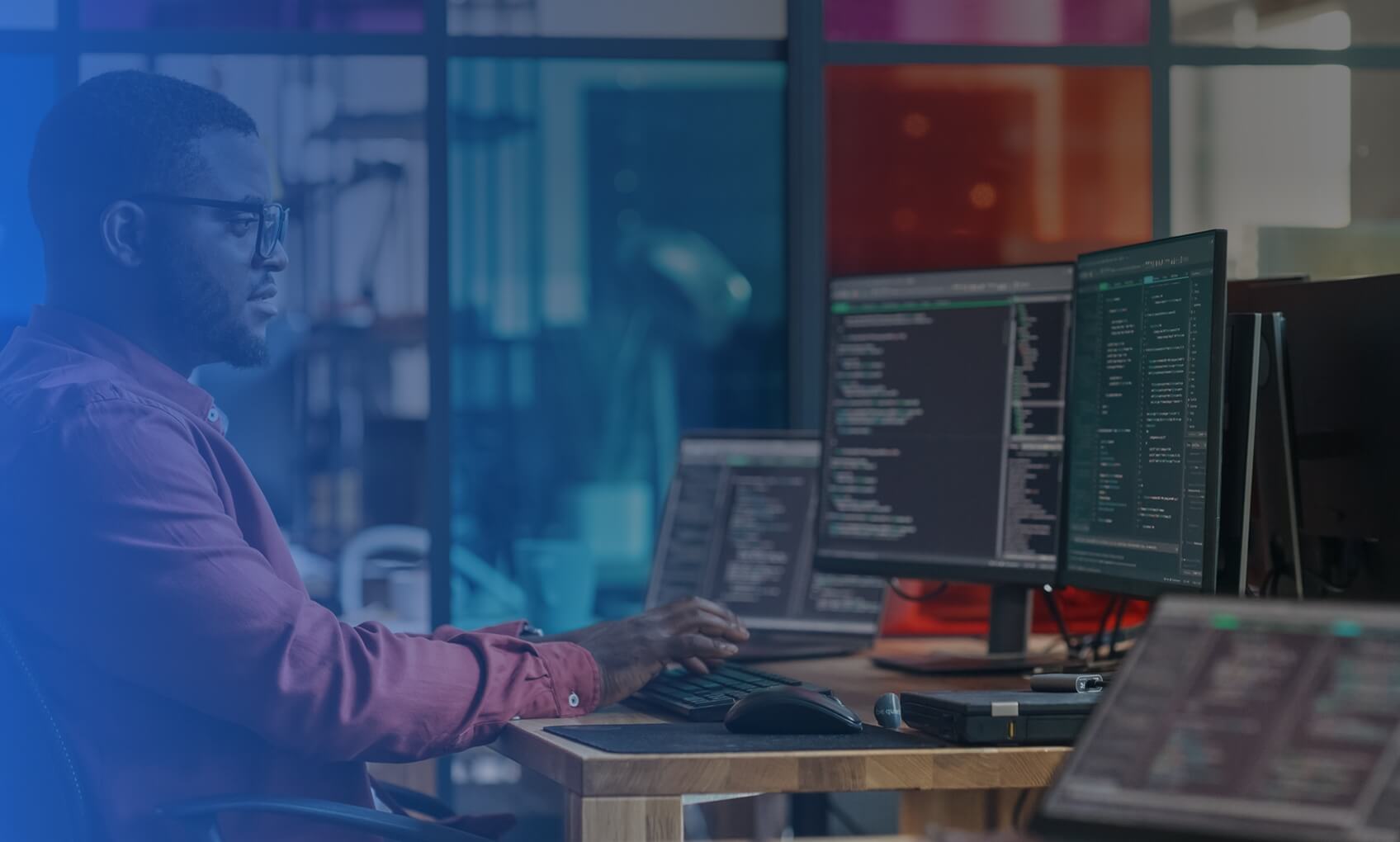 A person wearing glasses and a red shirt is sitting at a desk, typing on a keyboard and looking at multiple monitors displaying Azure Migration code in a modern office with colorful glass panels.