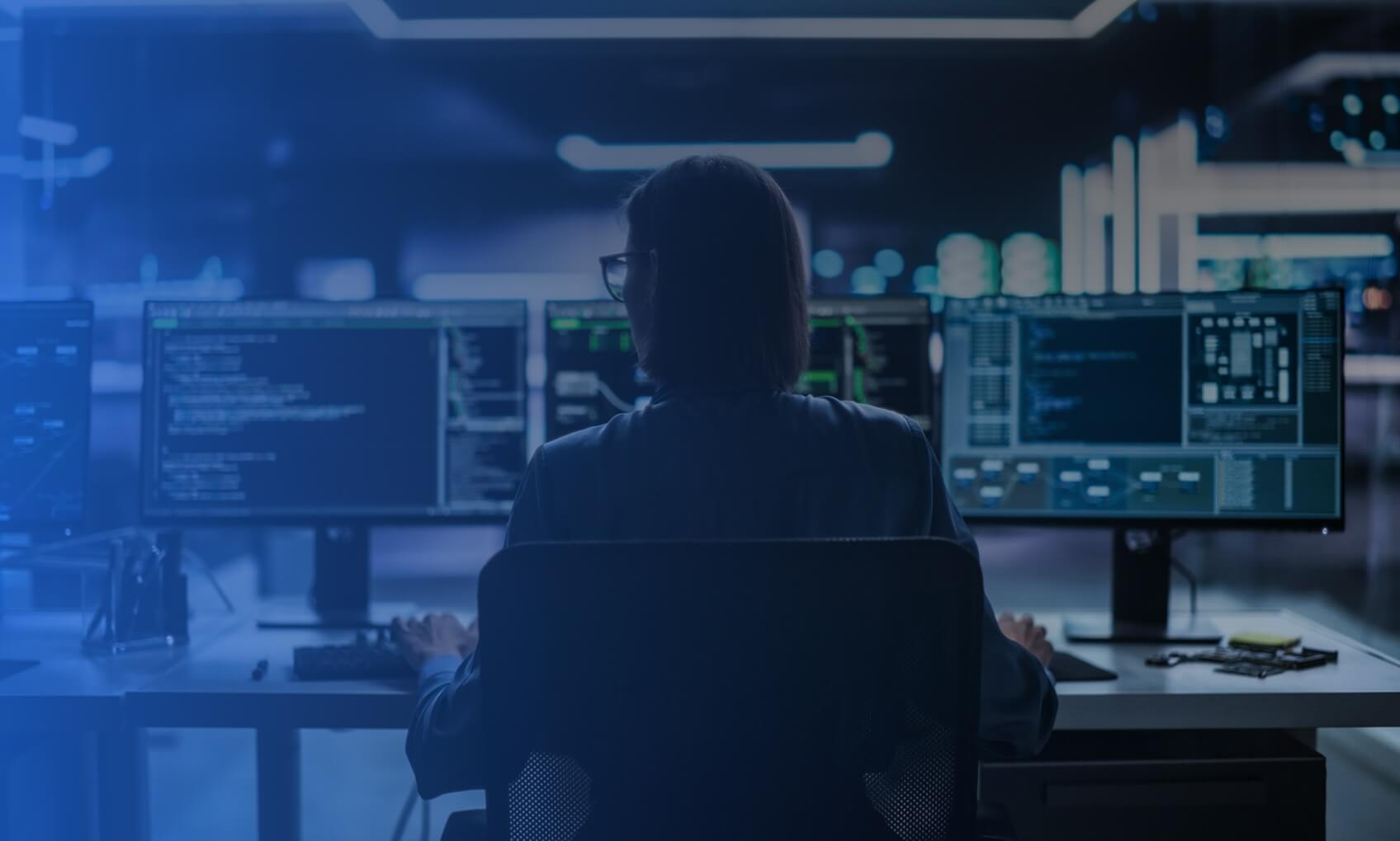 A person sits at a desk in a dimly lit room, facing multiple computer monitors displaying code and data, suggesting a programming or cybersecurity environment focused on Azure Migration.