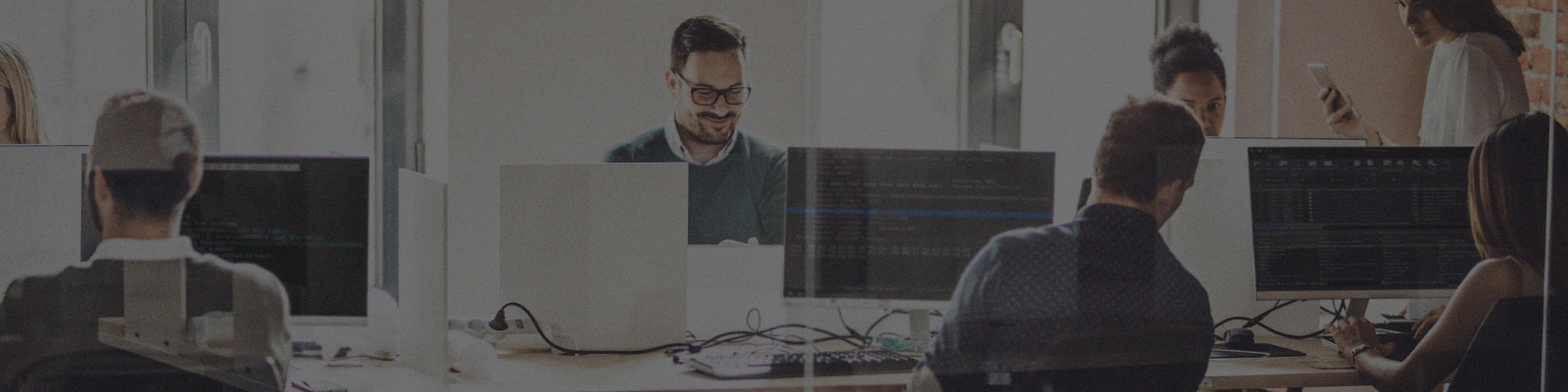 Several people work at desks with computers in a modern office, some focused on monitors while one person smiles, perhaps discussing an Azure Migration project. Large windows in the background fill the space with natural light.