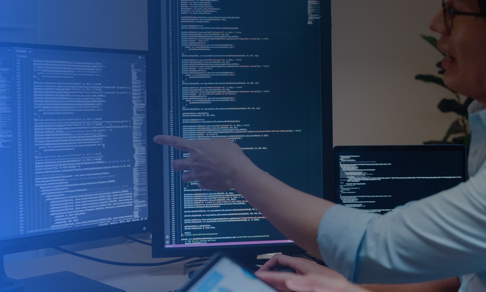 Two people look at computer monitors displaying lines of code. One points at the screen while the other observes, suggesting collaboration on an Azure Sentinel Implementation in an office setting.