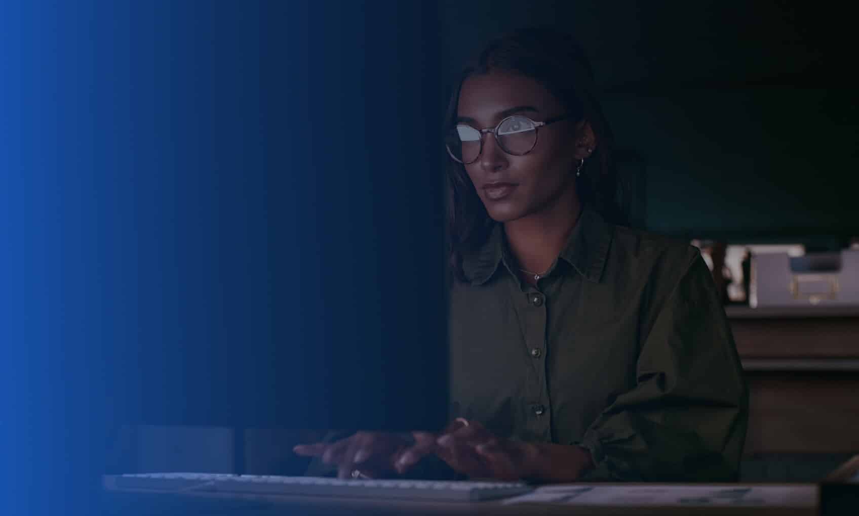 A woman wearing glasses and a dark green shirt works intently on Azure Sentinel Implementation in a dimly lit room, her face illuminated by the screen’s glow.