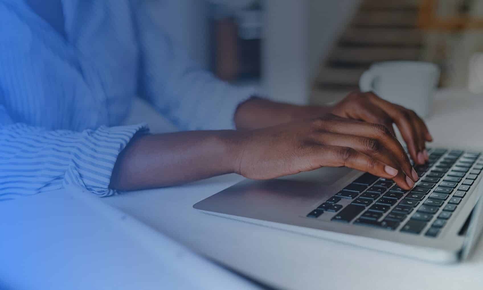 A person wearing a blue and white striped shirt types on a laptop keyboard at a white desk, possibly focused on Azure Sentinel Implementation, with a blurred coffee mug and stairs visible in the background.