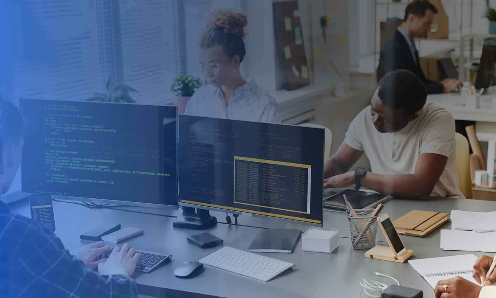 Four people work at desks with computers and papers in a bright, modern office. Multiple monitors display code and Azure Sentinel Implementation dashboards. Two people are focused on screens; others are writing or using a tablet.