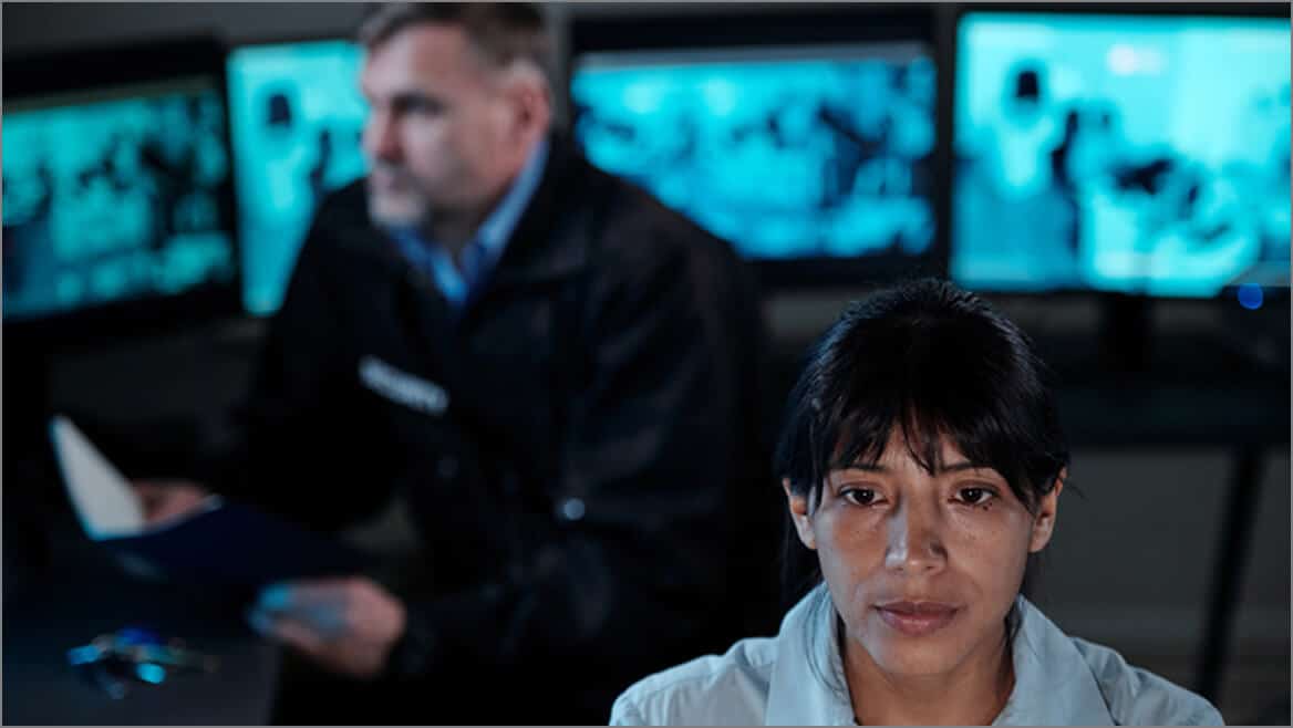A woman looks serious in the foreground while a man in a uniform sits behind her, holding a folder. Computer monitors with blurred images are visible in the background.