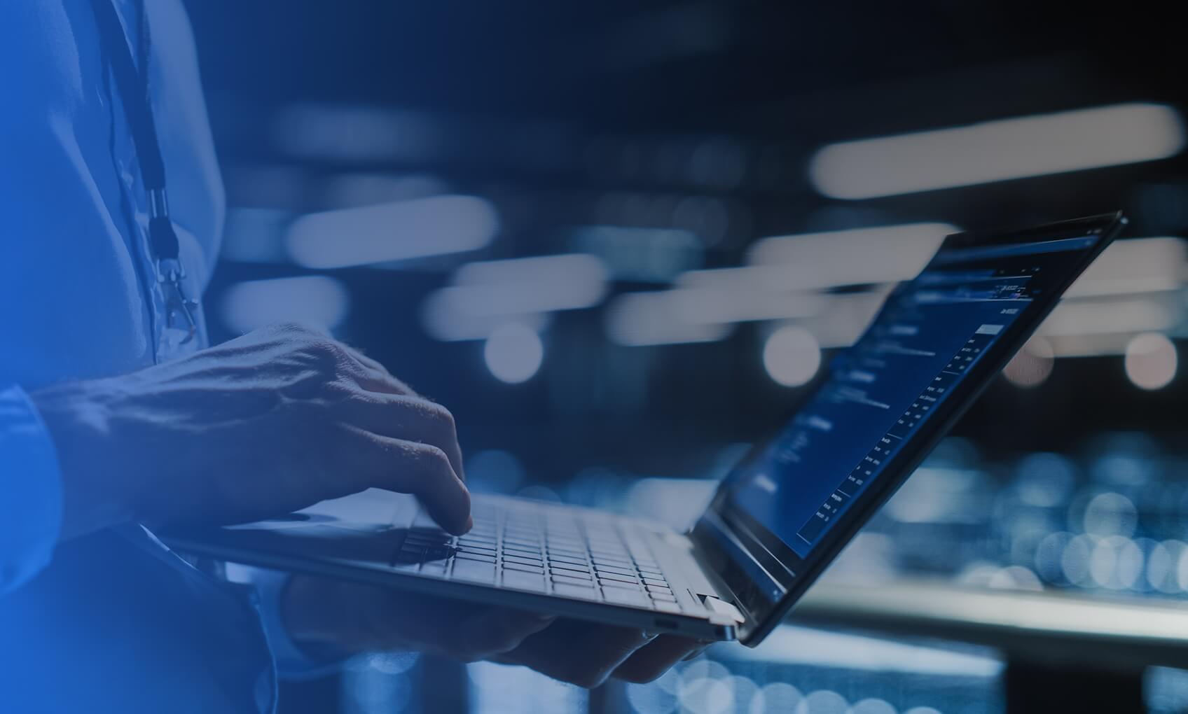 A person wearing a lanyard uses a laptop, typing with one hand. The blurred background and bright lights suggest a modern office where Email Security Implementation might be the focus.