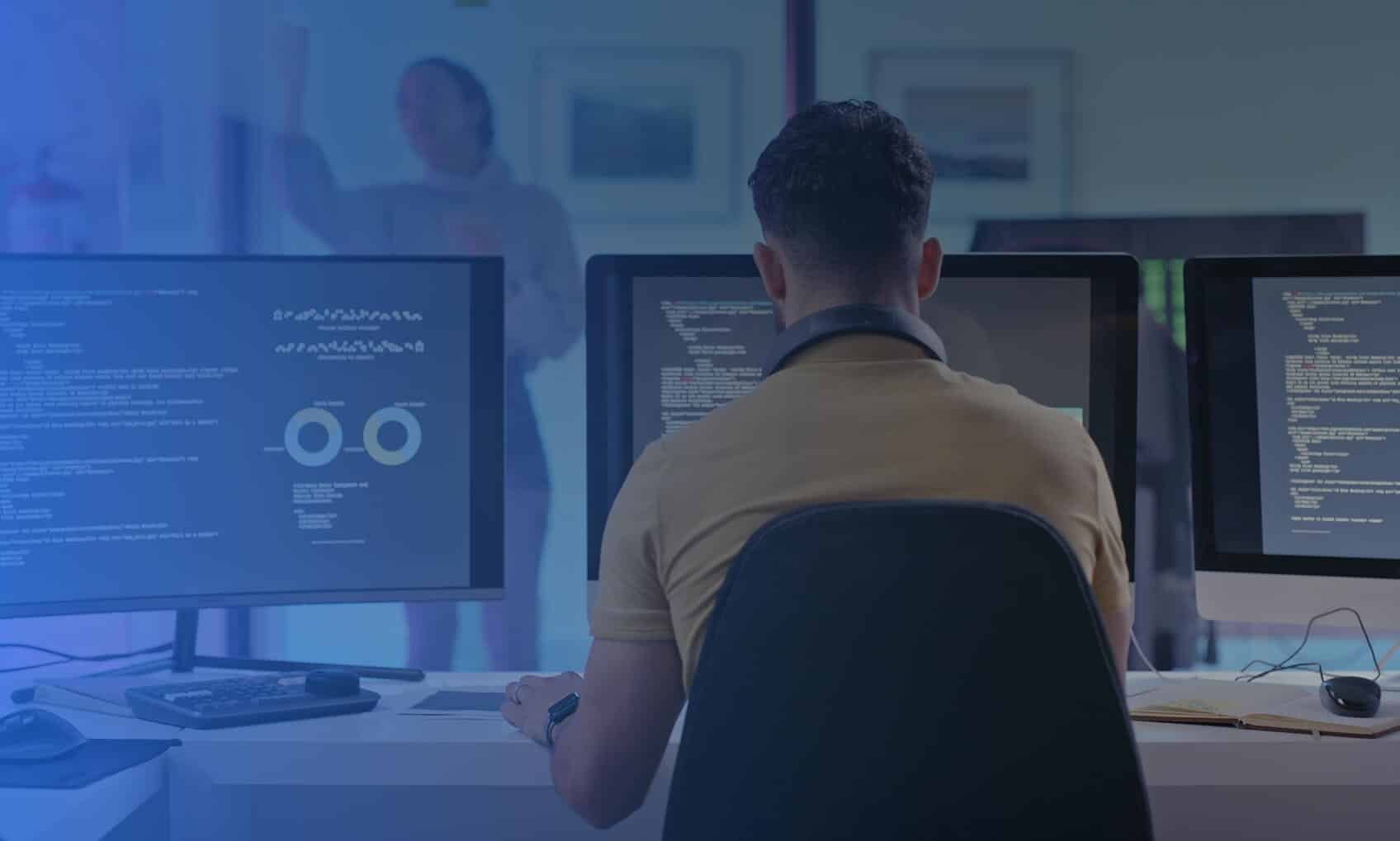 A person sits at a desk working on three computer monitors displaying code, data charts, and Email Security Implementation tasks, while another person stands in the background by a window.