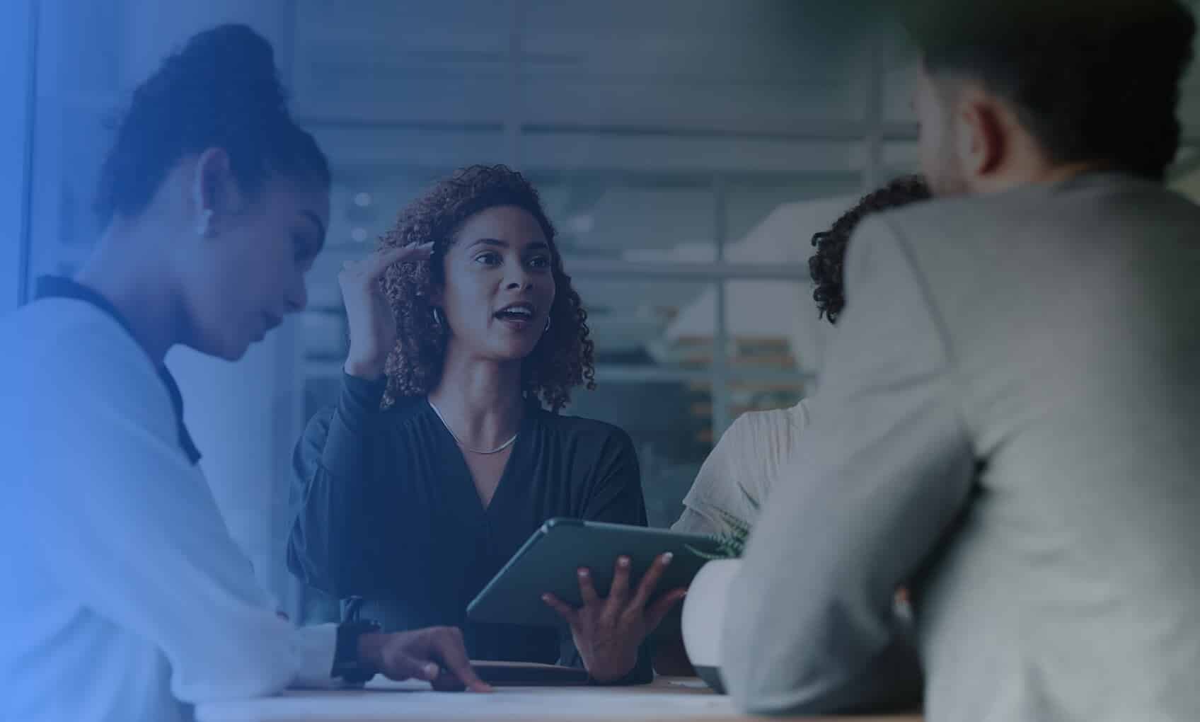 Three businesspeople sit around a table in a bright, modern office, engaged in discussion. The woman in the center gestures while speaking about Email Security Implementation, holding a digital tablet as the others listen attentively.