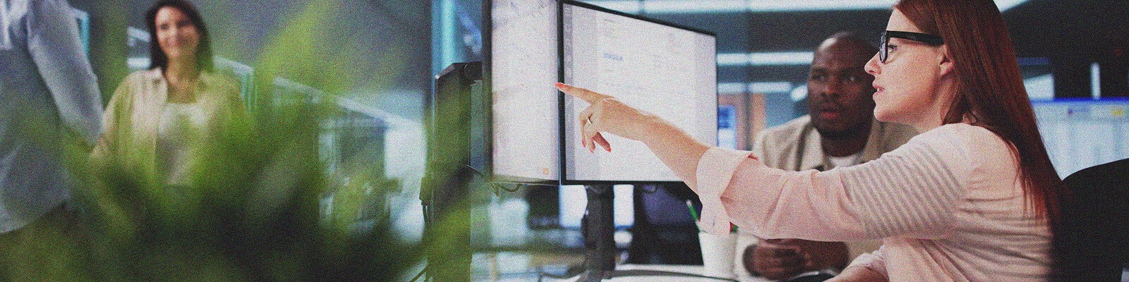 A woman wearing glasses points at a computer monitor, discussing Email Security Implementation with a male colleague in an office. Two others stand in the background, while a green plant is visible in the foreground.