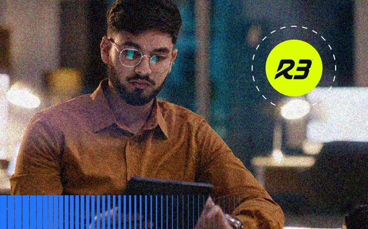 A man with glasses and a beard, wearing a brown shirt, sits at a desk in an office at night, looking at a tablet while researching GCC High Migration. Computer monitors and desk lamps are visible in the blurred background.