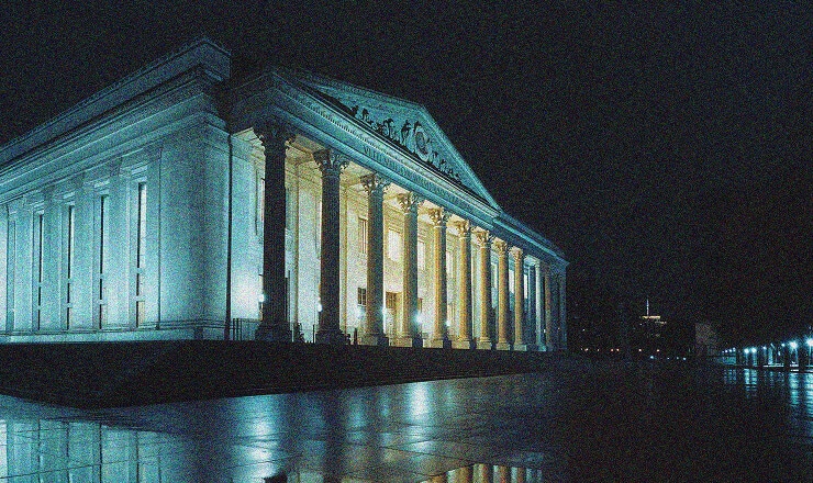 A grand, columned building is illuminated at night, reflecting off a wet surface. Reminiscent of classical architecture, its design echoes the strength and security upheld by FedRAMP Compliance. The sky is dark and the scene is quiet.