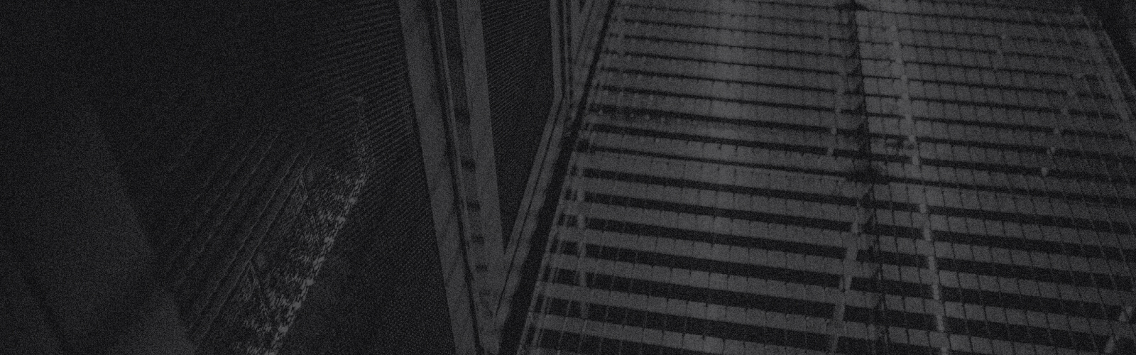 A dark, moody photo of a tall building with a grid-like pattern of windows, mostly in shadow. Diagonal lines and faint reflections echo an abstract, urban atmosphere—suggesting the quiet orderliness of FISMA Compliance.