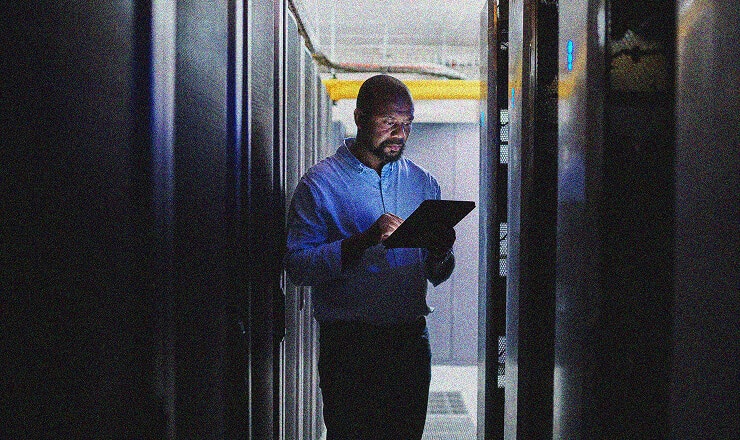 A man stands in a dimly lit server room, using a tablet while surrounded by tall racks of servers on both sides, ensuring CMMC Compliance.