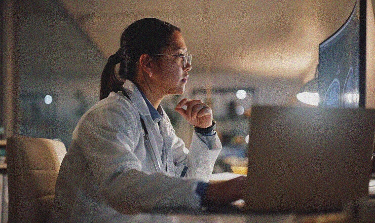 A doctor wearing a white coat and glasses looks intently at a computer screen in an office at night, ensuring HIPAA compliance while reviewing patient data, with a laptop and medical equipment nearby.