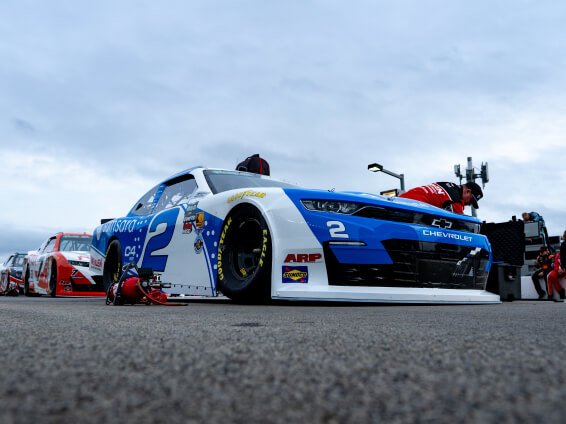 A blue and white Chevrolet race car with the number 2 on the side is parked on a racetrack. A team member in red works nearby, with another race car and equipment visible in the background under a cloudy sky.