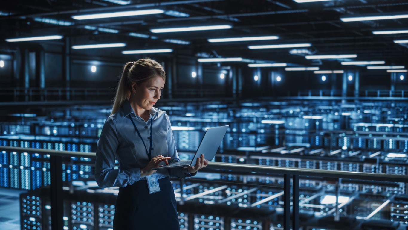 A woman in business attire stands in a large, modern data center, holding and working on a laptop. Rows of illuminated server racks fill the background, creating a high-tech environment.