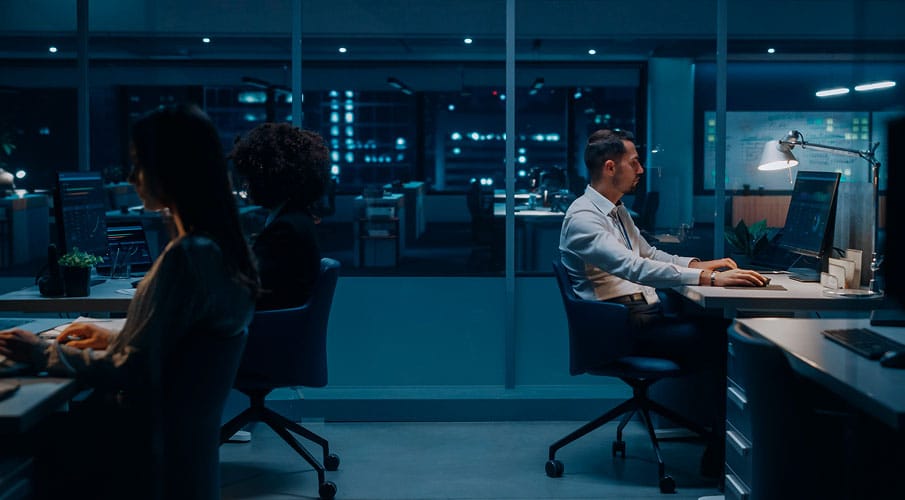 Three people work late at computers in a modern office, with city lights visible through large windows. The room is dimly lit, creating a focused, quiet atmosphere.