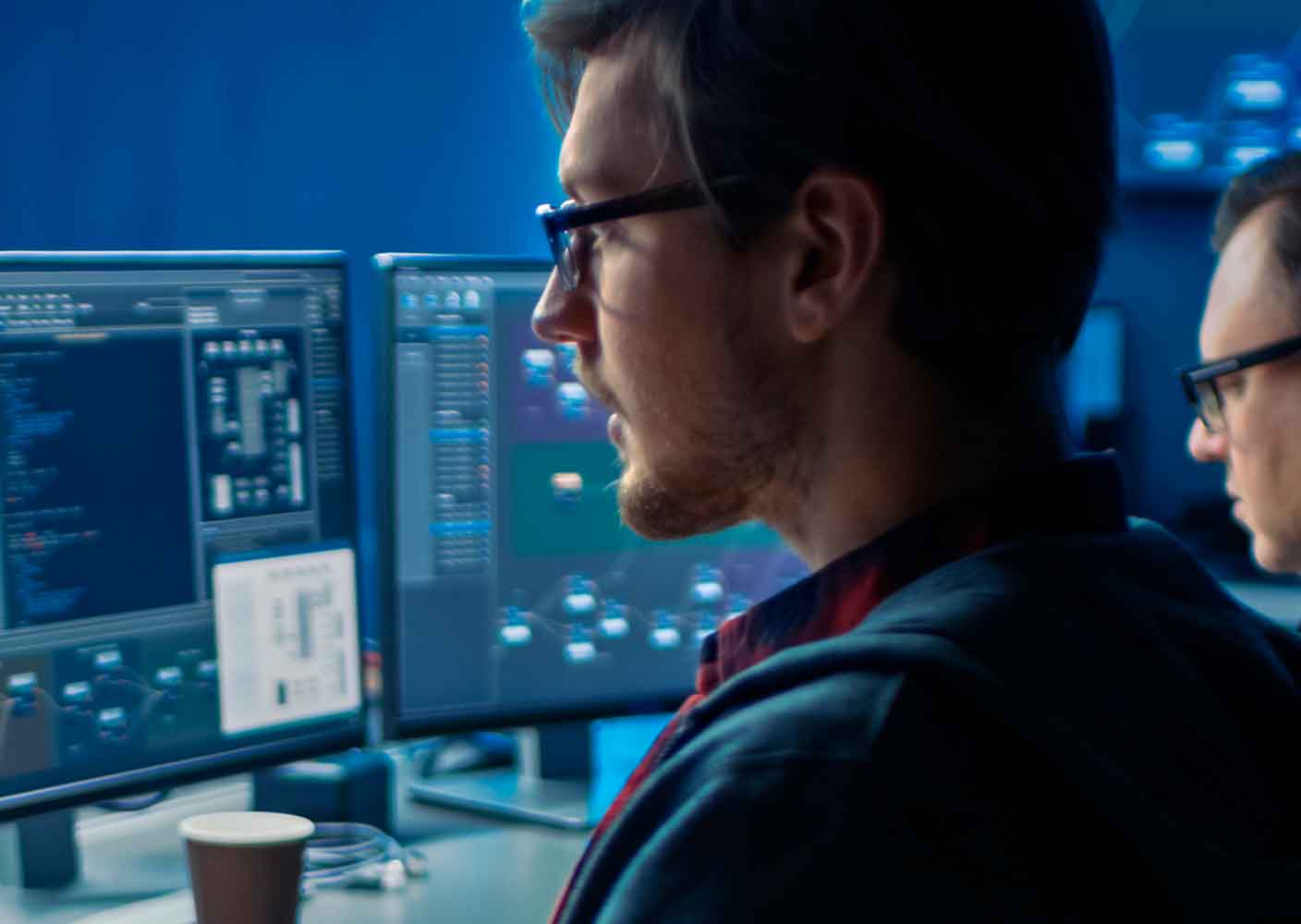 Two people work at computer desks with multiple monitors displaying code and software interfaces in a dimly lit office, with a coffee cup on one desk in the foreground.