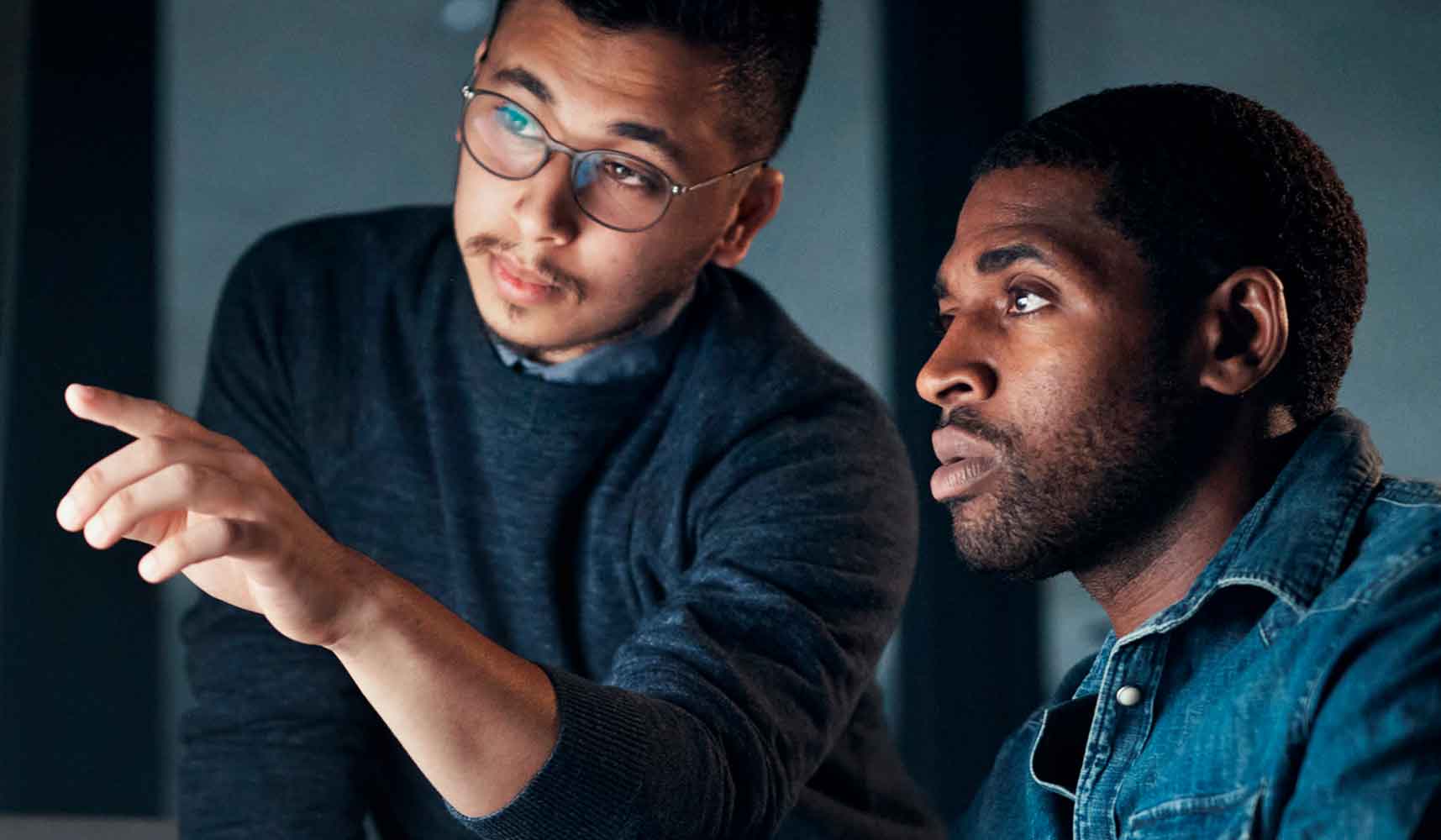 Two men sit closely together, focusing intently on something off-screen. One man, wearing glasses, is pointing while explaining something to the other, who listens attentively. Both appear to be working or collaborating.