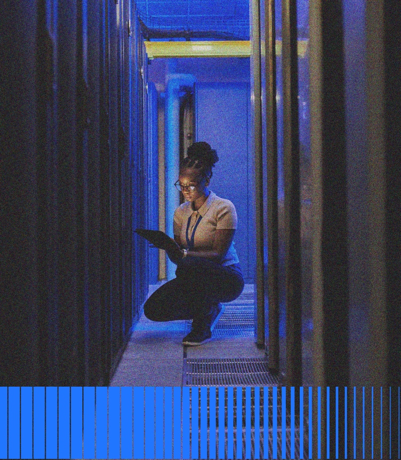 A woman kneels in a dimly lit server room, illuminated by blue lights, while working on a tablet device between rows of server racks.
