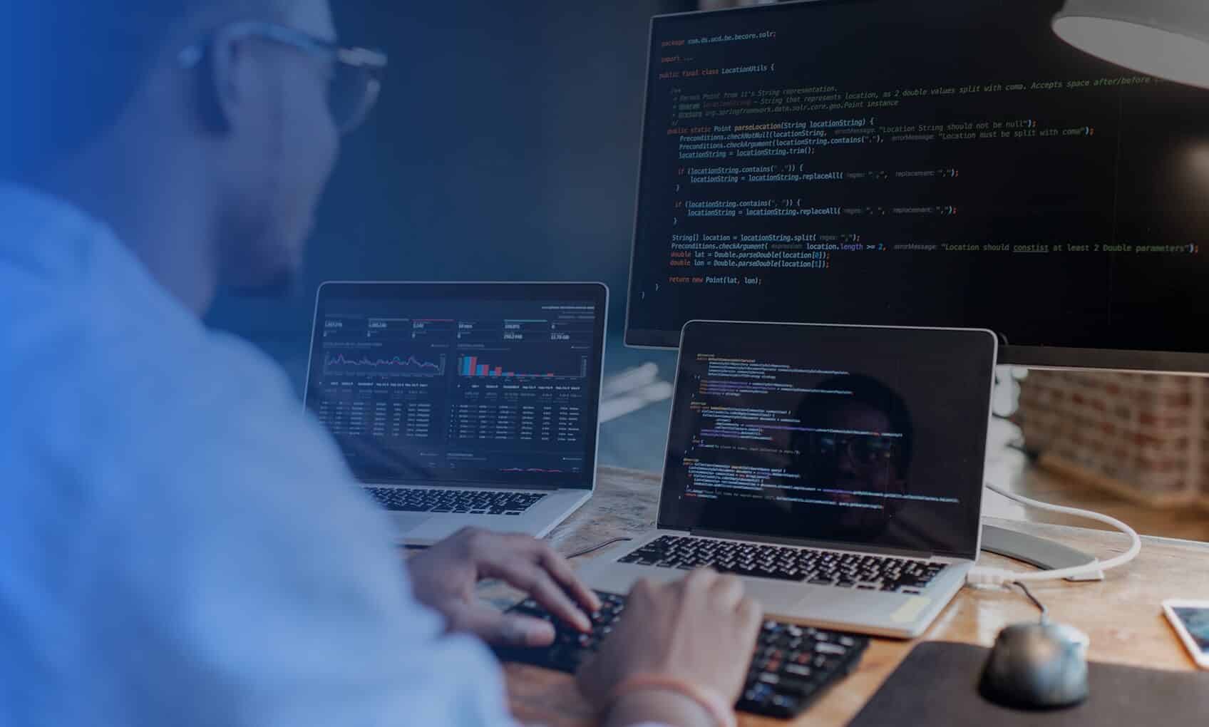 A person sits at a desk working on two laptops with code and data displayed, focusing on Intune and Entra-ID Implementation, while a large monitor in the background shows lines of programming code.