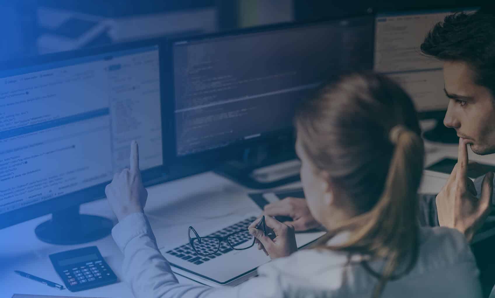 Two people sit at a desk with multiple computer monitors, discussing Intune and Entra-ID implementation. One points at the monitor while the other observes thoughtfully. A calculator and notebook are on the desk.