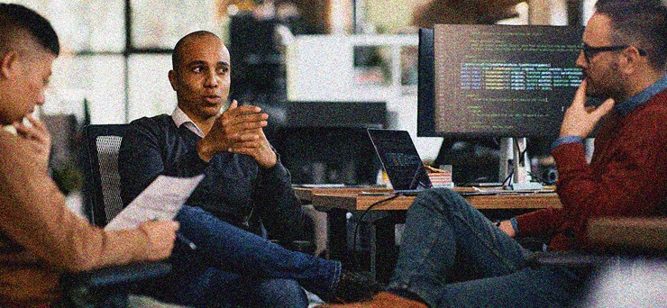 Three people sit in an office discussing Intune and Entra-ID implementation. One gestures while speaking, another holds papers, and the third listens thoughtfully. Computer monitors with code are visible in the background.