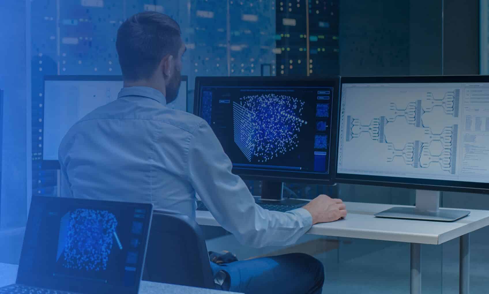 A man sits at a desk in an office, working on multiple computer monitors displaying 3D data visualizations, neural network diagrams, and Intune and Entra-ID Implementation dashboards, with large windows showing a cityscape at night.