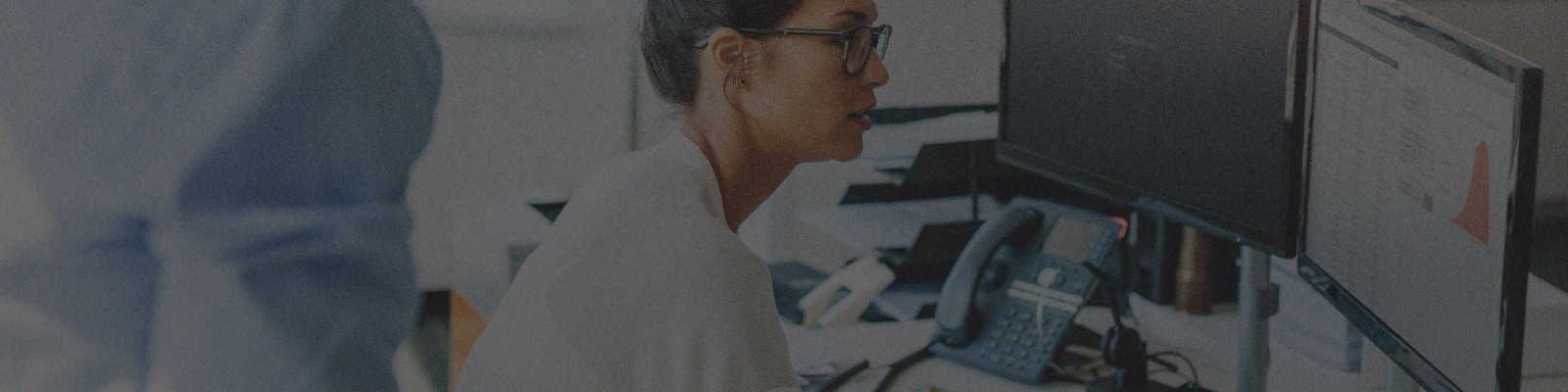 A woman wearing glasses works at a desk with dual computer monitors, a phone, and documents, while another person stands nearby, partially visible.
