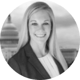 A woman with long blonde hair, wearing a blazer and blouse, smiles at the camera. The background shows the U.S. Capitol building, suggesting a professional or political setting. The image is in black and white.