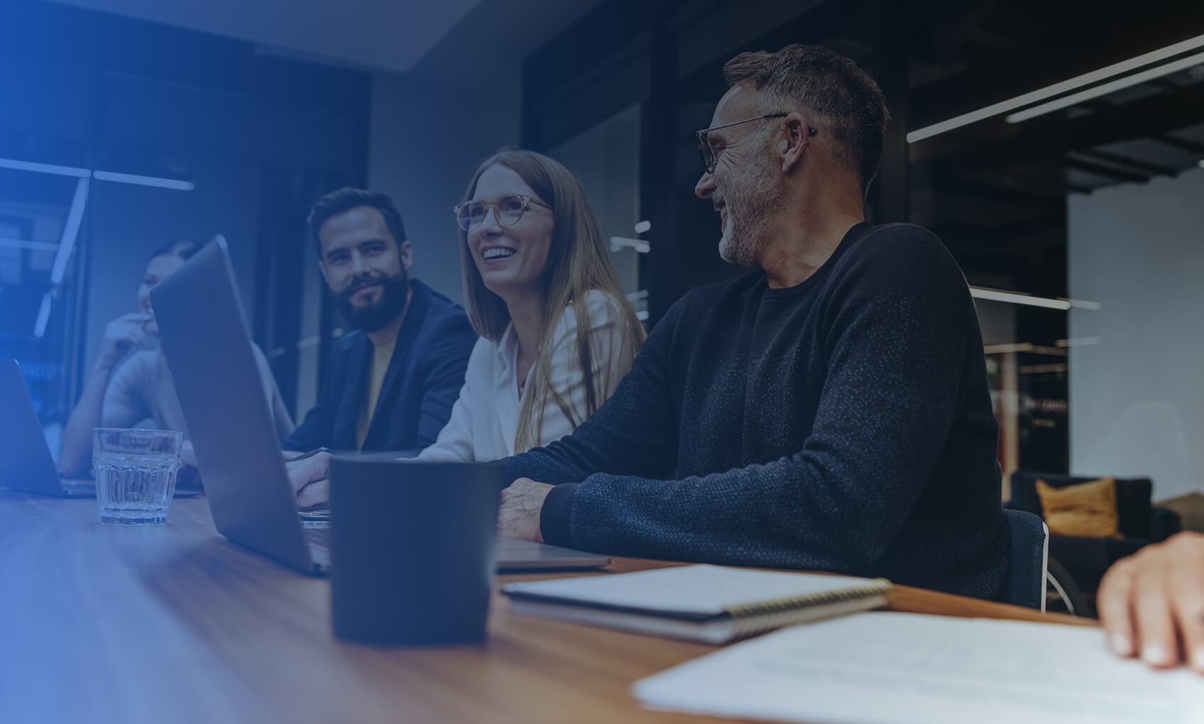 Three people sitting indoors, appearing engaged and smiling; the person in focus is waving and wearing glasses, while two others, possibly discussing M365 Tenant Consolidation, are slightly out of focus and seated nearby.