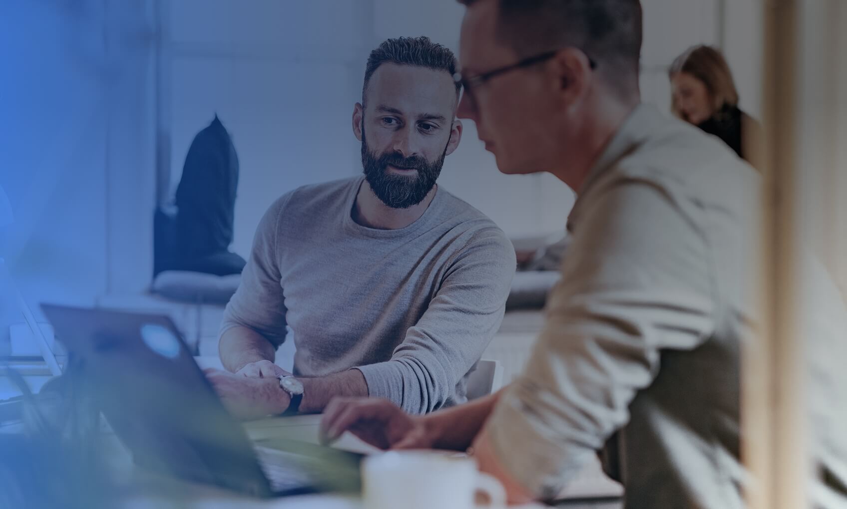 Two men are sitting at a desk, working together on laptops in a bright office, discussing M365 Tenant Consolidation. One man listens attentively while the other speaks, with a woman blurred in the background.