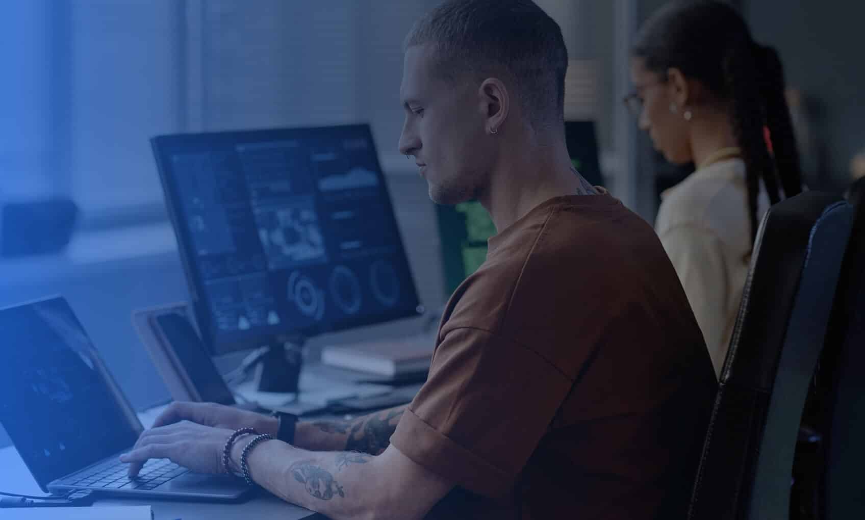 A man types on a laptop at a desk, with large monitors displaying M365 Tenant Consolidation data in front of him. A woman sits at a nearby workstation, focusing on her own screen. The office appears modern and well-lit.