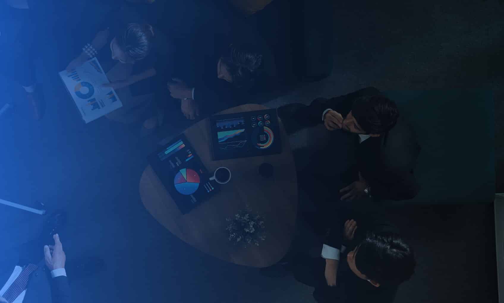 A group of businesspeople sit around a small table discussing M365 Tenant Consolidation, reviewing charts and graphs on digital tablets and documents, some holding coffee cups as they analyze colorful data.