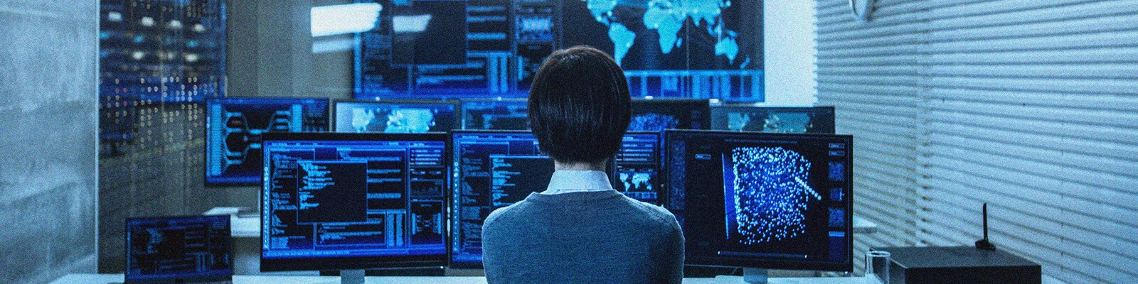 A person with dark hair in a bun, seen from behind, sits at a desk surrounded by multiple monitors displaying data and maps, focused on M365 Tenant Consolidation in a high-tech control room.