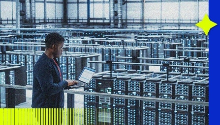 A person stands on a balcony overlooking a large data center filled with rows of server racks, typing on a laptop. Blue lights illuminate the CloudOps teams spacious, industrial environment.