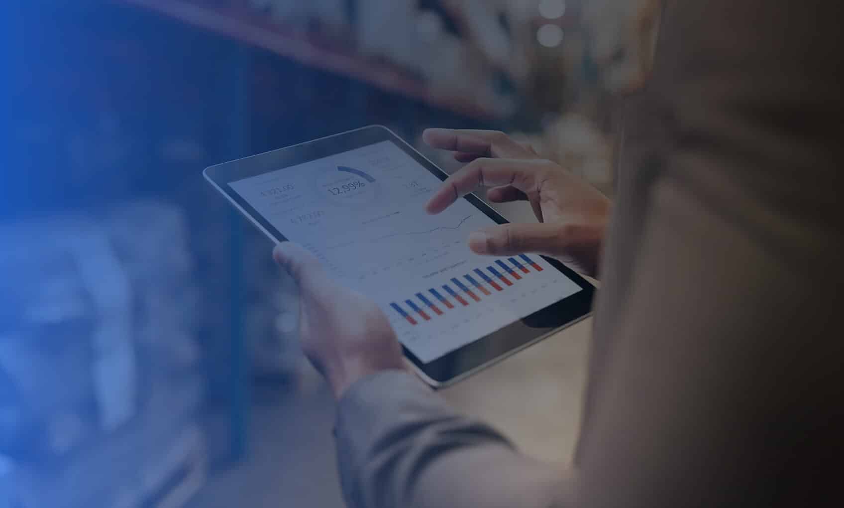 A person holding a tablet displays a CloudOps data analytics dashboard with charts and graphs, likely in a warehouse setting, as blurred shelves and inventory are visible in the background.