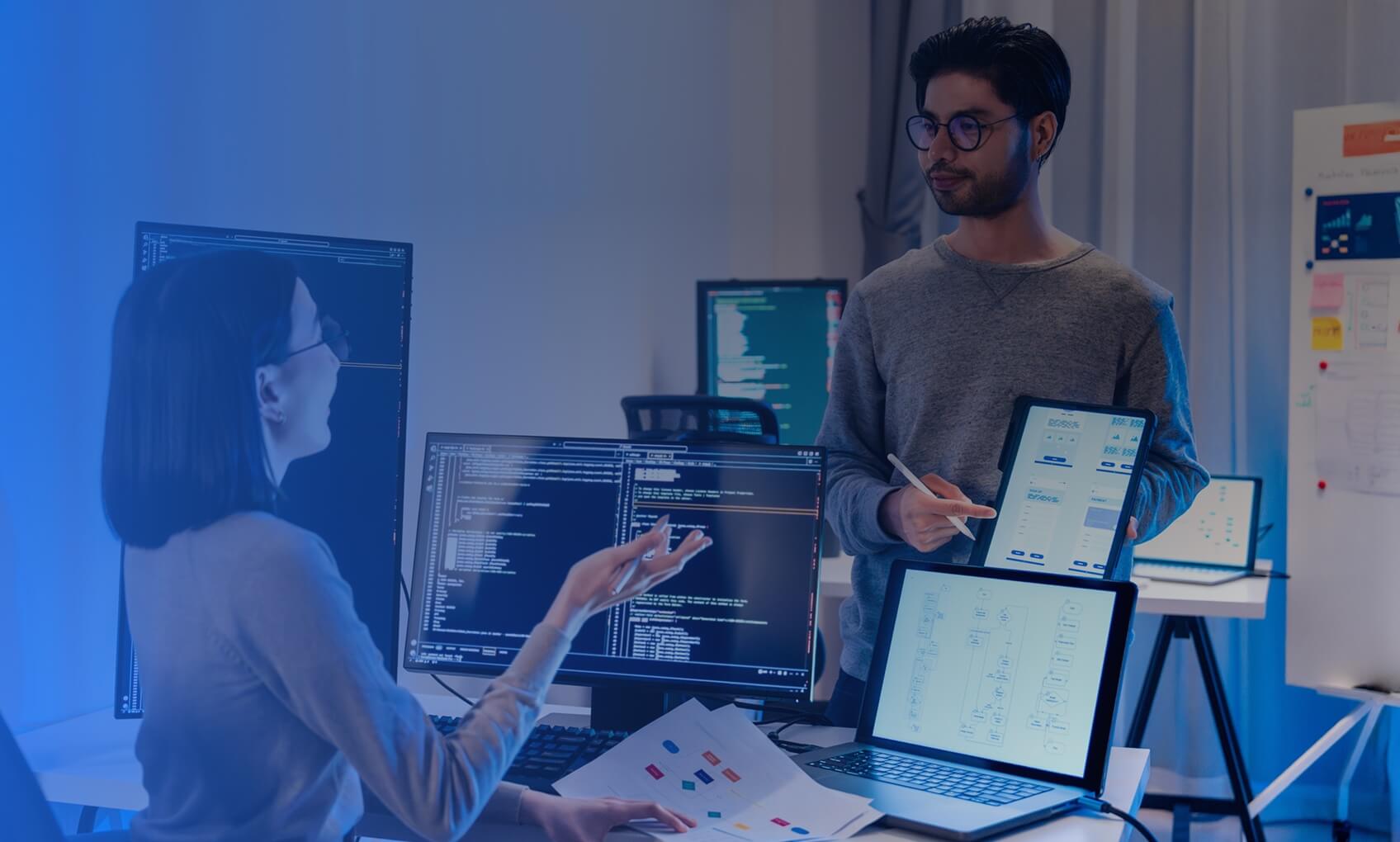 Two people in an office discuss code on computer monitors; one is seated and gesturing, the other stands holding a tablet displaying charts, highlighting the benefits of dedicated IT services amid screens and whiteboards in the background.