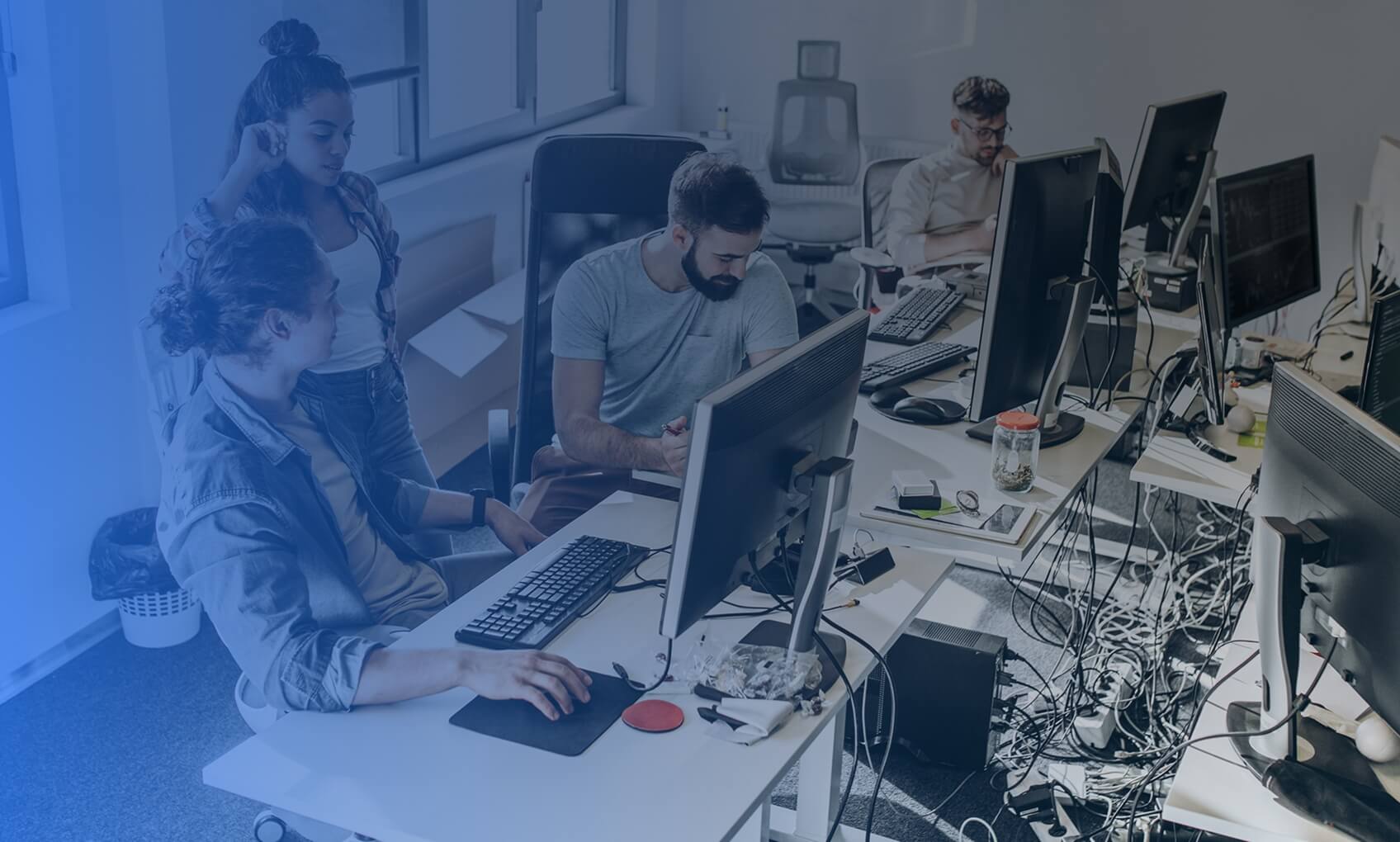 Four people work at desks with computers in a modern office. Two collaborate while looking at a monitor, showcasing dedicated IT services, while another types on a keyboard and one works alone. The office is bright with large windows.