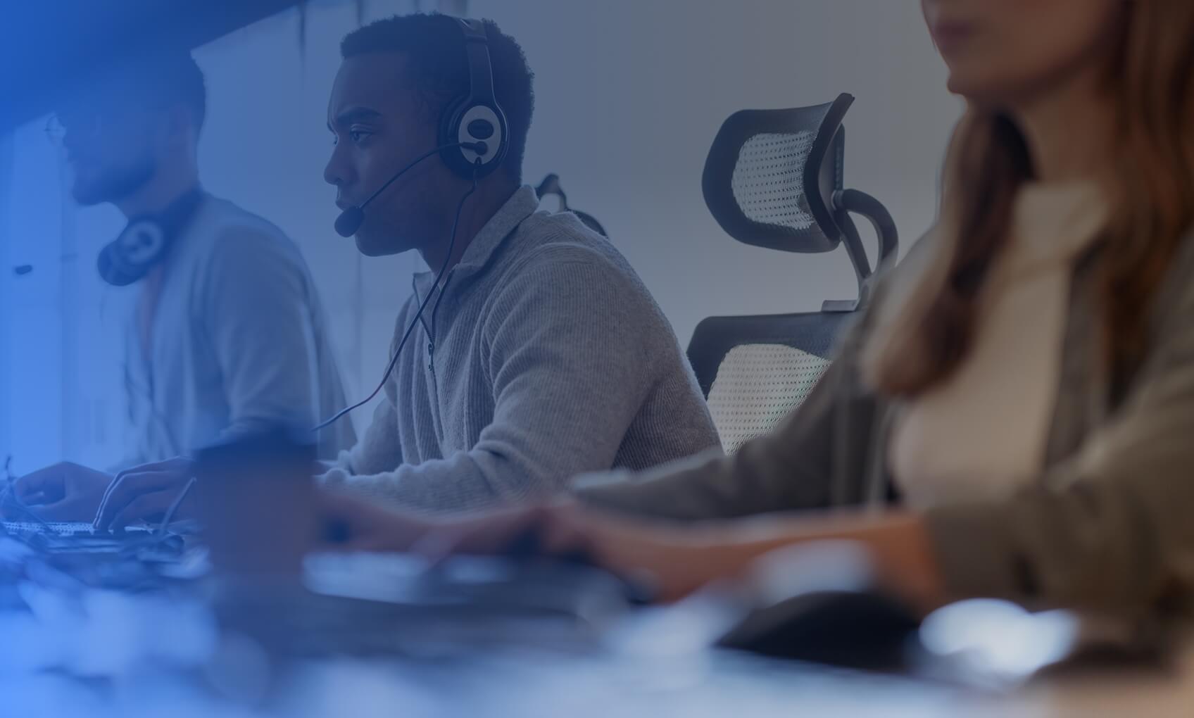 Three people wearing headsets sit at desks with computers in a bright office, focusing on their work. The scene suggests a customer support or call center environment specializing in dedicated IT services.