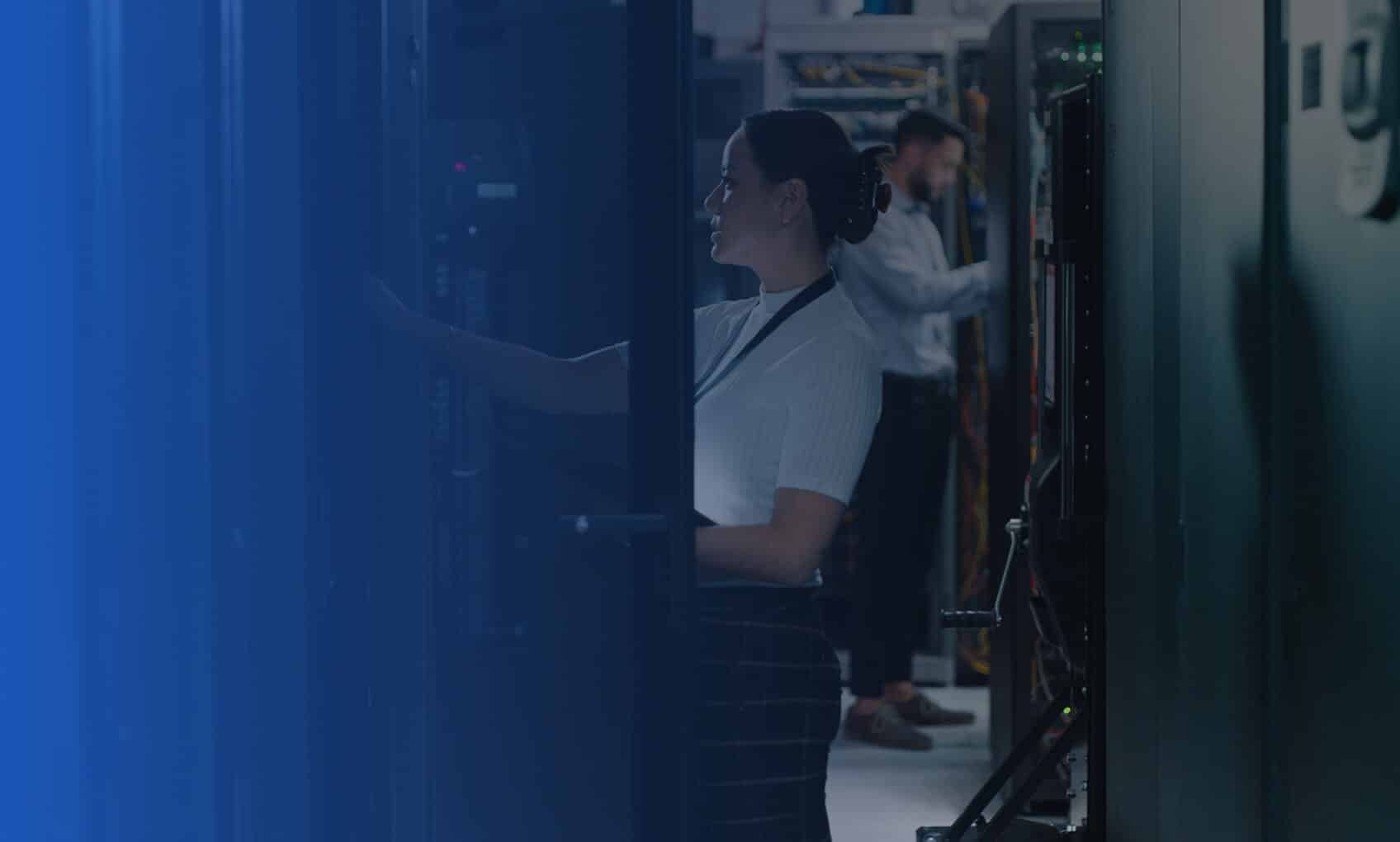 Two people in uniforms work inside a server room surrounded by tall racks of computer equipment, demonstrating the precision and focus that come with dedicated IT services. One checks a server in the foreground while another works in the background.
