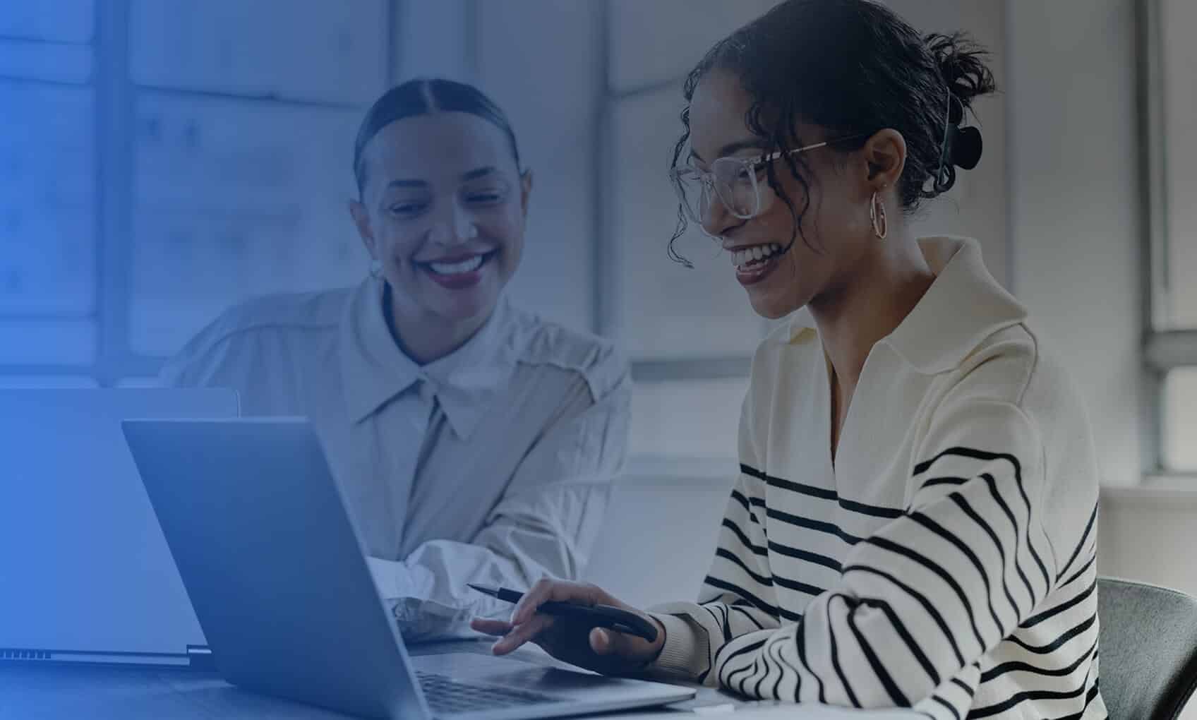 Two women smiling and working together at a laptop in a bright, modern office, reflecting the collaborative spirit behind dedicated IT services. Large windows fill the workspace with natural light.