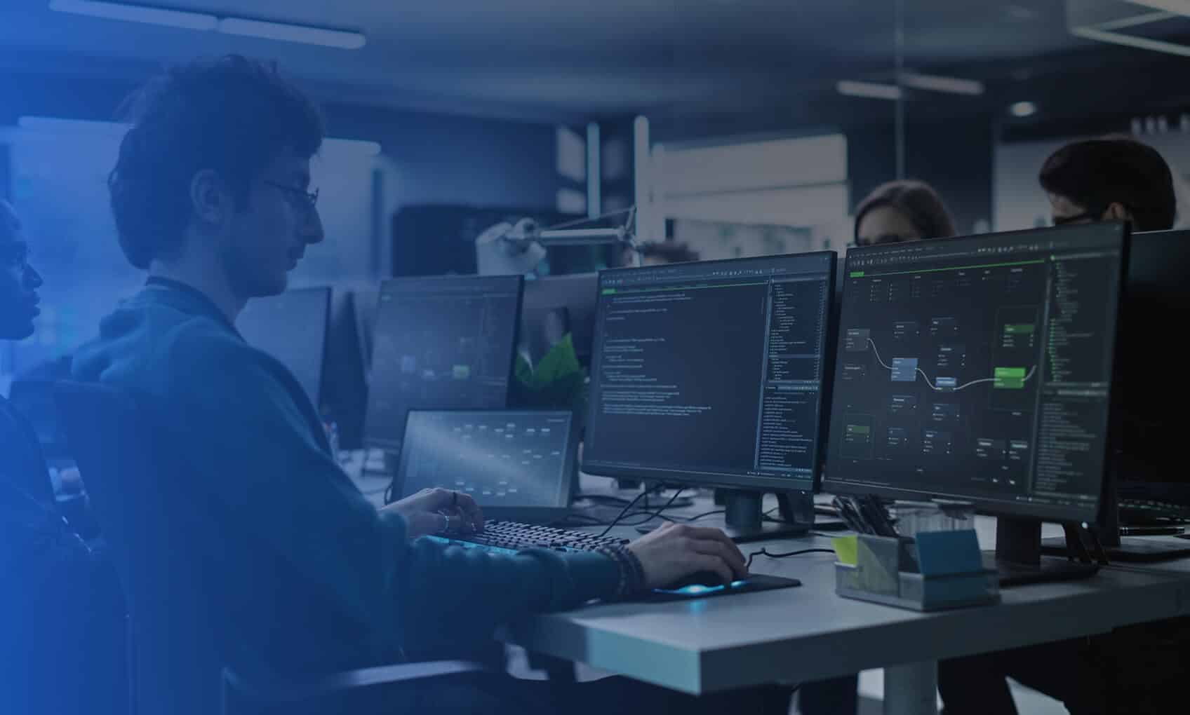 A person works at a desk with three large monitors displaying code and flowcharts in the R3 Network Operations Center (NOC), surrounded by other people and workstations. The lighting is dim and focused, creating a modern, high-tech atmosphere.