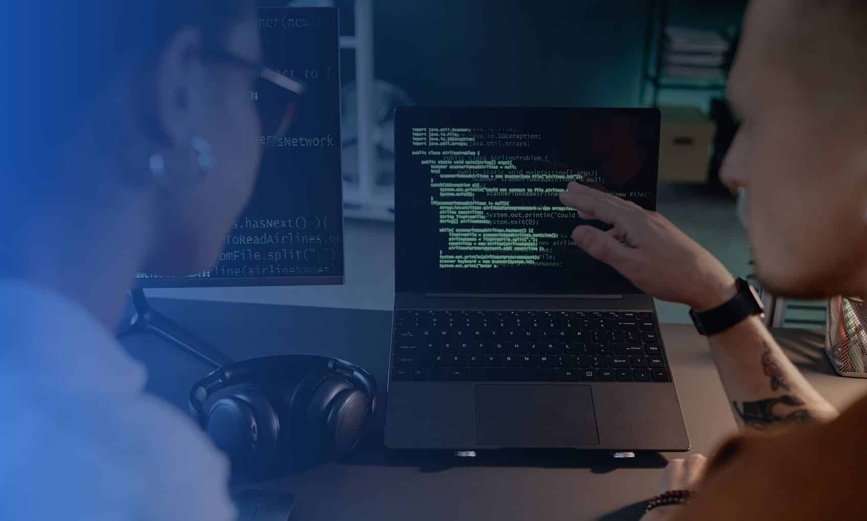 Two people sit at a desk in the R3 Network Operations Center (NOC), reviewing lines of code on a laptop screen. One points at the display while headphones and an additional monitor with code are also visible on the desk.